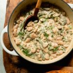 An overhead shot shows a braiser pot filled with creamy dilly beans and peas. A serving spoon sticks out of the pot and some bread is seen to the side.
