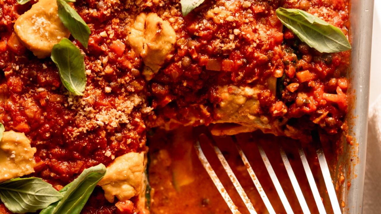 An up close, overhead shot shows a baked vegan lasagna in a baking dish. The top is finished with little dollops of tofu cashew ricotta, fresh basil leaves and vegan