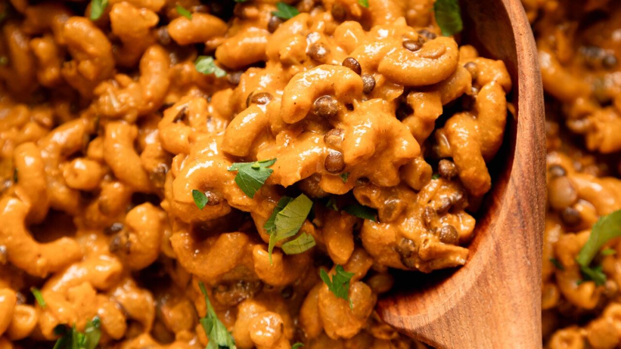 An up close, overhead shot shows a vegan hamburger helper-style mixture with lentils, elbow macaroni, and chopped parsley. A wooden spoon is sticking out of the mixture.