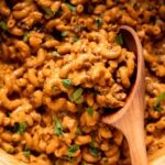 An up close, overhead shot shows a vegan hamburger helper-style mixture with lentils, elbow macaroni, and chopped parsley. A wooden spoon is sticking out of the mixture.